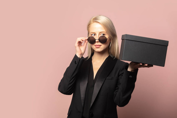 Стильная блондинка в блейзере и солнцезащитных очках с черной подарочной коробкой на розовом фоне - Фото, изображение
