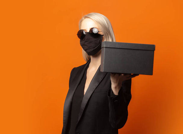 Estilo rubio en blazer negro con caja de regalo sobre un exuberante fondo naranja - Foto, imagen