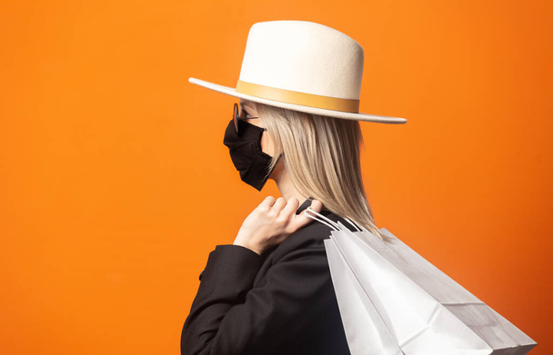 Стильная блондинка в черном блейзере и белой шляпе с пакетами для покупок на пышном оранжевом фоне - Фото, изображение