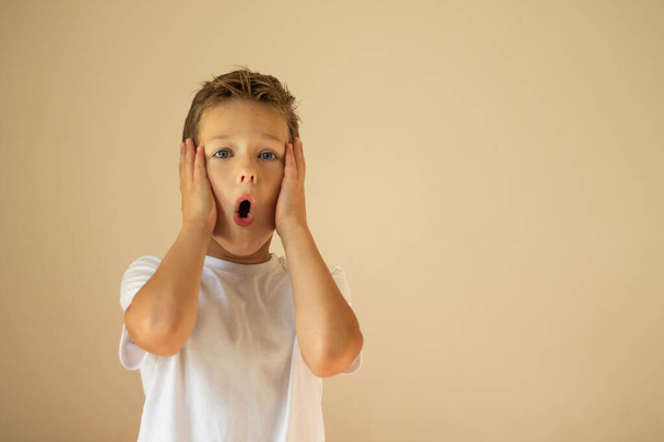 Un garçon surpris ou effrayé de 7 à 10 ans dans un T-shirt blanc se tient debout et crie avec ses mains sur ses joues sur un fond beige. Espace de copie. - Photo, image