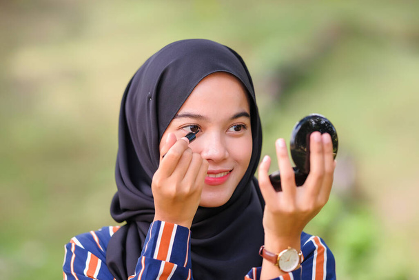 Красивая мусульманка в черном хиджабе наносит макияж с помощью подводки для глаз и смотрит в зеркало - Фото, изображение