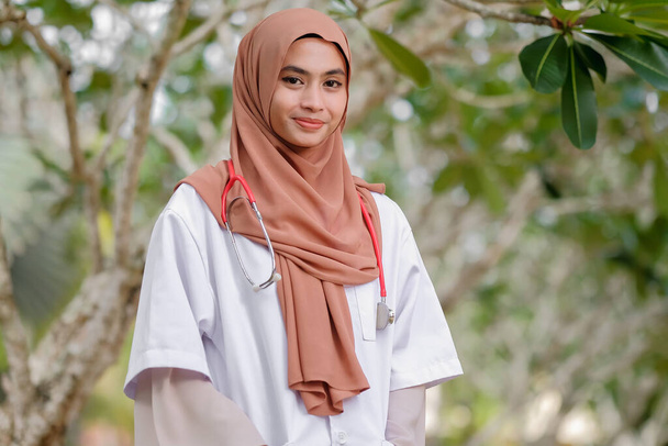 Joven prethy médico de la ASEAN con hijab y estetoscopio en el jardín del hospital. Concepto de servicio médico y sanitario. - Foto, imagen