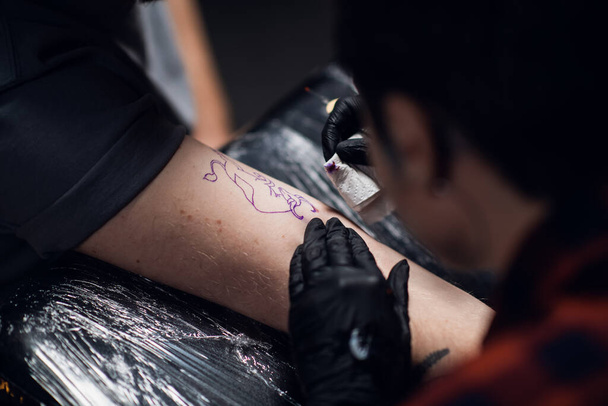 Foto de cerca de una artista de tatuajes que pone un tatuaje en el brazo de un joven en un salón de tatuajes usando el método de mano. - Foto, Imagen