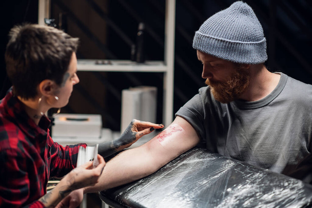 Un tatuatore benda la mano dei giovani uomini con un sottile strato di gelatina di petrolio dopo aver applicato il tatuaggio. - Foto, immagini