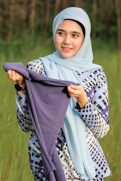 Porträt der glücklichen schönen jungen Model in modischen Hijab-Stil posiert im grünen Bereich, während sie Stoff hält. Stilvolles muslimisches Hijab-Mode-Lifestyle-Porträtkonzept. - Foto, Bild