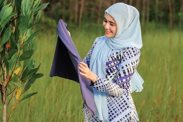 Retrato de modelo joven hermosa feliz en estilo hijab de moda posando en el área verde mientras sostiene la tela. Estilo musulmán femenino hijab moda estilo de vida retrato concepto. - Foto, imagen