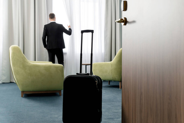 Rear view of businessman wearing dark suit standing in hotel room looking into window - 写真・画像