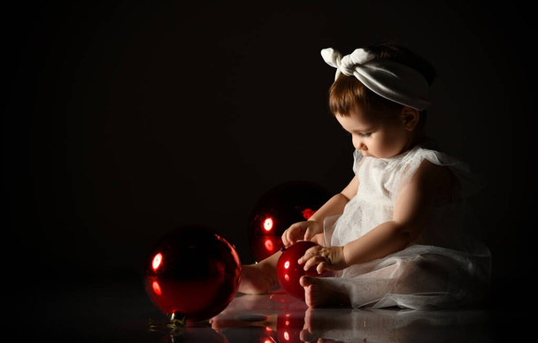 Little child in white headband and dress, barefoot. She playing with two red balls, sitting on floor. Twilight, black background. - Φωτογραφία, εικόνα