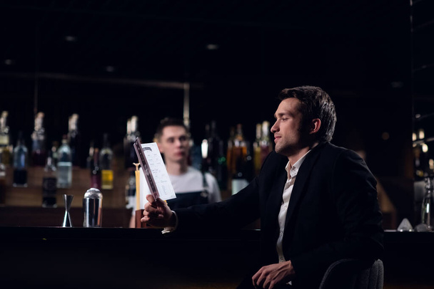 χαριτωμένος άντρας διαβάζει το μενού του μπαρ, κάθεται στον πάγκο - Φωτογραφία, εικόνα