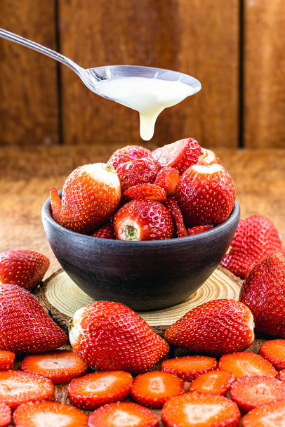 βάζο από φρέσκια φράουλα με συμπυκνωμένο γάλα (ξινή κρέμα) που στάζει πάνω από τα φρούτα, ξύλινο φόντο με χώρο για κείμενο - Φωτογραφία, εικόνα