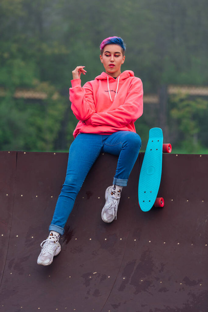 Portrait of a trendy pretty young girl with short colored hair and nose piercing dressed in pink hoodie and jeans sitting next to the skateboard coart with her blue plastic skateboard. - Photo, image