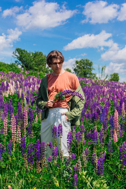 Tall handsome man in a green jacket standing on lupine flowers field holding lupine flowers in hand, enjoing the beauty of nature. Man surrounded by purple and pink lupines. - Foto, imagen