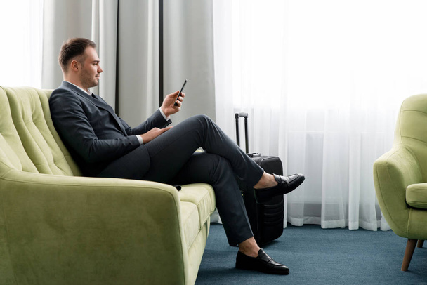 Wesoły biznesmen korzystający z telefonu siedzącego w pokoju hotelowym z walizką w podróży służbowej - Zdjęcie, obraz