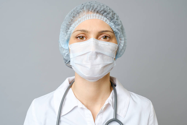 Portrait of young female doctor in protective medical mask on her face and cap on her head. Isolated on gray background - Photo, Image