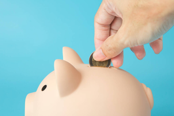 Mano sosteniendo una moneda, alcancía de cerdo sobre fondo azul para el concepto de ahorro de dinero - Foto, imagen