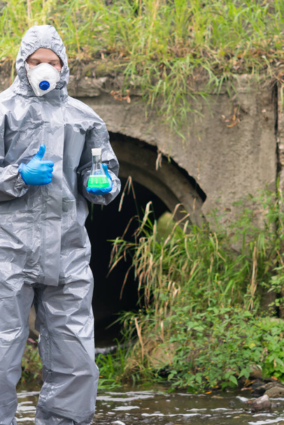 a man in a protective suit and mask holds a flask of green liquid in his hands and shows his thumb up on the background of sewage pipes coming out from the factory - Фото, изображение