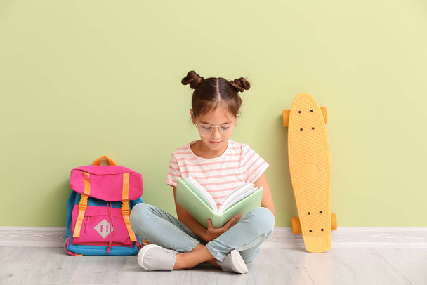 Klein schoolmeisje met skateboard en notebook zitten in de buurt van kleur muur - Foto, afbeelding