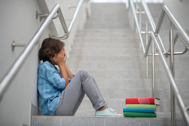 Schoolboy sitting with knees bent on stairs, covering his face - Foto, Imagen