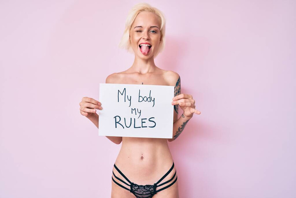 Jeune femme blonde avec tatouage debout torse nu tenant bannière avec mon corps mes règles message coller la langue heureux avec une expression drôle.  - Photo, image