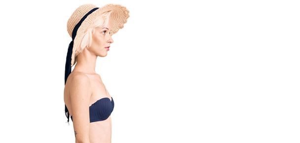 Jeune femme blonde avec un tatouage portant un bikini et un chapeau d'été regardant vers le côté, pose de profil relax avec un visage naturel avec un sourire confiant.  - Photo, image