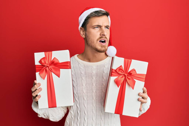 Handsome caucasian man wearing christmas hat and holding gifts angry and mad screaming frustrated and furious, shouting with anger. rage and aggressive concept.  - 写真・画像