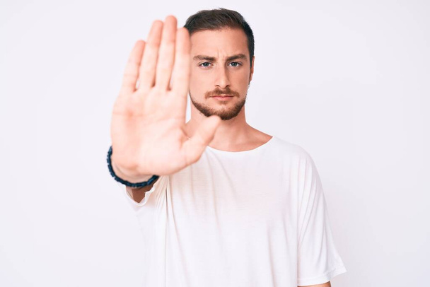 Jonge knappe man met een casual wit T-shirt die niet meer zingt met de palm van de hand. waarschuwingsuitdrukking met negatief en ernstig gebaar op het gezicht.  - Foto, afbeelding