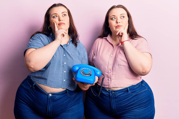 Young plus size twins holding vintage telephone serious face thinking about question with hand on chin, thoughtful about confusing idea  - Foto, Bild