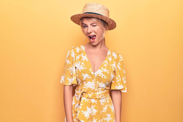 Όμορφη ξανθιά γυναίκα σε διακοπές φορώντας καλοκαιρινό καπέλο και φόρεμα πάνω από κίτρινο φόντο κλείνει το μάτι κοιτάζοντας την κάμερα με σέξι έκφραση, χαρούμενο και χαρούμενο πρόσωπο. - Φωτογραφία, εικόνα