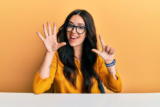 Krásná brunetka mladá žena nosí brýle a ležérní oblečení sedí na stole ukazuje a ukazuje prsty číslo sedm a zároveň se usmívá sebevědomě a šťastně.  - Fotografie, Obrázek