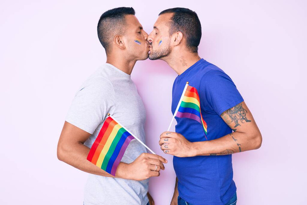 Young couple of gay men together in love holding homosexual pride colorful flag kissing with affection - Photo, Image