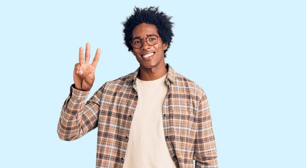 bel homme afro-américain aux cheveux afro portant des vêtements décontractés et des lunettes montrant et pointant vers le haut avec les doigts numéro trois tout en souriant confiant et heureux.  - Photo, image