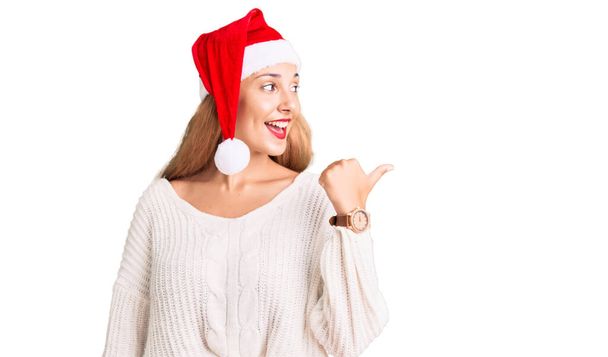 Bella giovane donna che indossa il cappello di Natale sorridente con volto felice guardando e indicando il lato con pollice in su.  - Foto, immagini