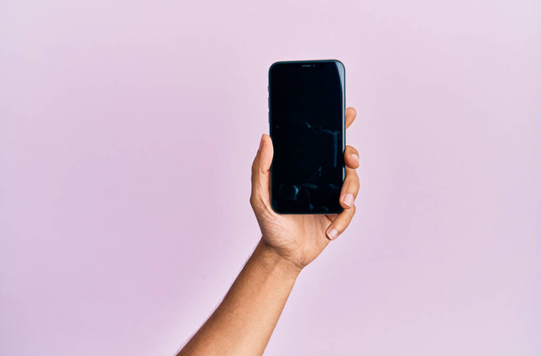 Dłoń młodego Latynosa pokazuje smartfona na odizolowanym różowym tle. - Zdjęcie, obraz