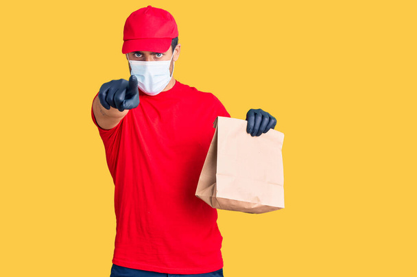 Young hispanic man delivering food wearing covid-19 safety mask holding paper bag pointing with finger to the camera and to you, confident gesture looking serious  - Valokuva, kuva