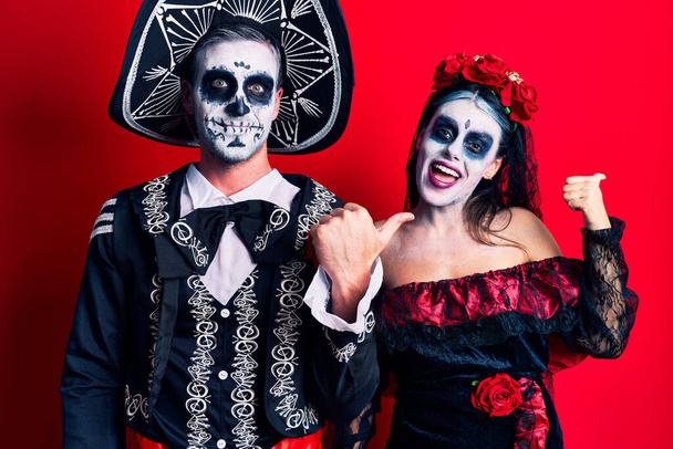 Young couple wearing mexican day of the dead costume over red smiling with happy face looking and pointing to the side with thumb up.  - Photo, Image