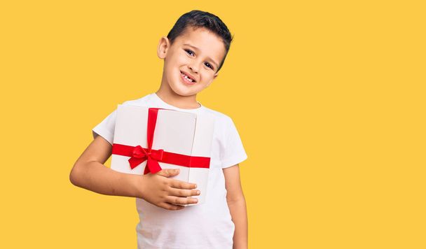 Little cute boy kid holding gift looking positive and happy standing and smiling with a confident smile showing teeth  - Фото, изображение