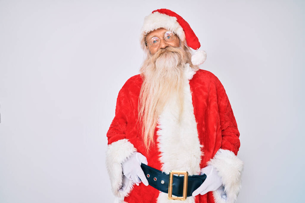 Γέρος ηλικιωμένος άνδρας με γκρίζα μαλλιά και μακριά γενειάδα φορώντας κοστούμι Santa Claus αναζητούν θετική και ευτυχισμένη στέκεται και χαμογελά με αυτοπεποίθηση χαμόγελο που δείχνει τα δόντια  - Φωτογραφία, εικόνα