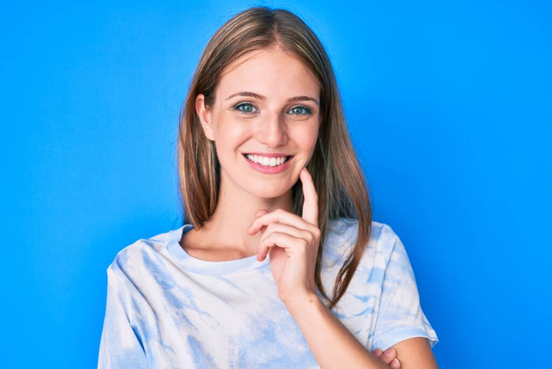 Young blonde girl wearing casual clothes smiling looking confident at the camera with crossed arms and hand on chin. thinking positive.  - Foto, imagen