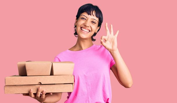 Young brunette woman with short hair holding take away food doing ok sign with fingers, smiling friendly gesturing excellent symbol  - Photo, Image
