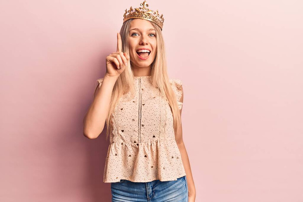 Jonge blonde vrouw draagt prinses kroon wijzen vinger omhoog met een succesvol idee. opgewonden en gelukkig. Nummer een.  - Foto, afbeelding
