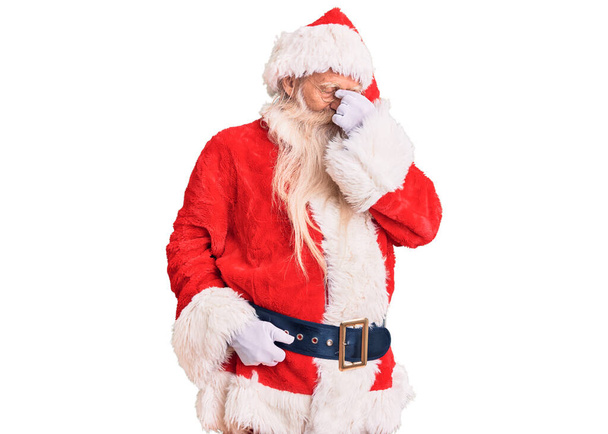 Старий старший чоловік з сірим волоссям і довгою бородою в традиційному костюмі Санта-Клауса втомився терти ніс і очі відчувають втому і головний біль. концепція стресу та розчарування
.  - Фото, зображення