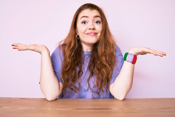 Young redhead woman wearing casual clothes sitting on the table clueless and confused with open arms, no idea and doubtful face.  - Photo, Image