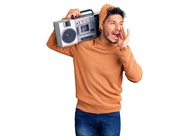 Handsome latin american young man holding boombox, listening to music shouting and screaming loud to side with hand on mouth. communication concept.  - Photo, Image