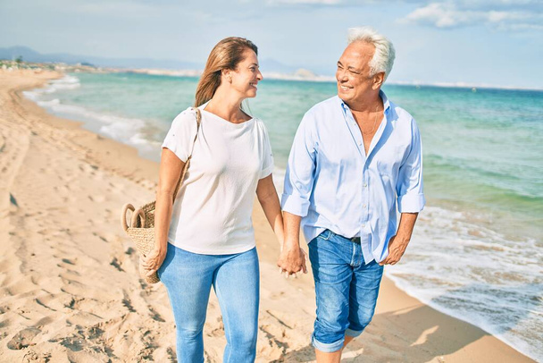 Middelbare leeftijd Latijns-Amerikaans paar glimlachend gelukkig wandelen op het strand. - Foto, afbeelding