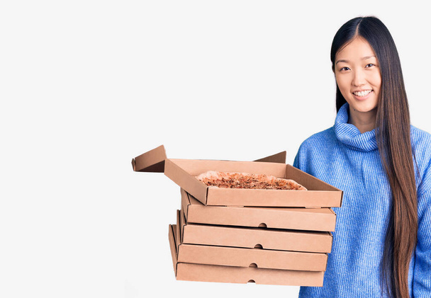 Mladá krásná čínská žena drží lepenkové krabice italské pizzy vypadá pozitivně a šťastně stojící a usmívá se s sebevědomým úsměvem ukazující zuby  - Fotografie, Obrázek