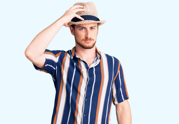 Young handsome caucasian man wearing summer hat confuse and wonder about question. uncertain with doubt, thinking with hand on head. pensive concept.  - Foto, Imagen