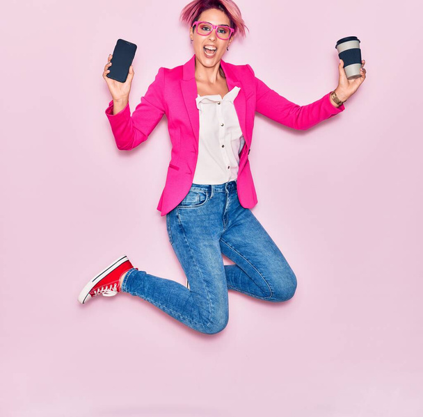 Young beautiful businesswoman with pink short hair wearing glasses smiling happy. Jumping with smile on face holding smartphone drinking coffee over isolated background - Photo, Image