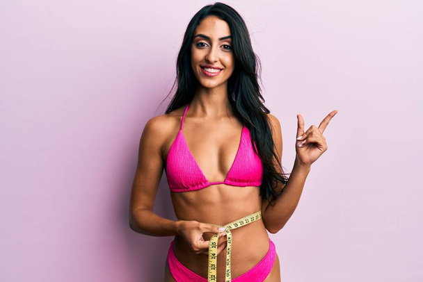Bella donna ispanica indossando bikini e misurando la vita sorridente felice indicando con mano e dito di lato  - Foto, immagini