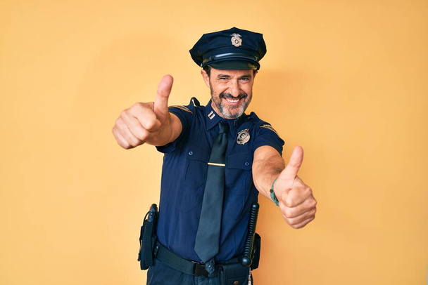 Latynos w średnim wieku, noszący mundur policyjny, aprobujący pozytywne gesty ręką, uśmiechający się z uśmiechem na ustach i cieszący się sukcesem. gest zwycięzcy.  - Zdjęcie, obraz