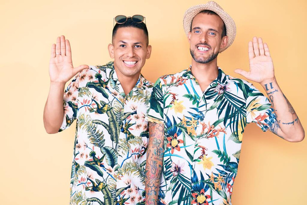 Young gay couple of two men wearing summer hat and hawaiian shirt waiving saying hello happy and smiling, friendly welcome gesture  - Photo, Image
